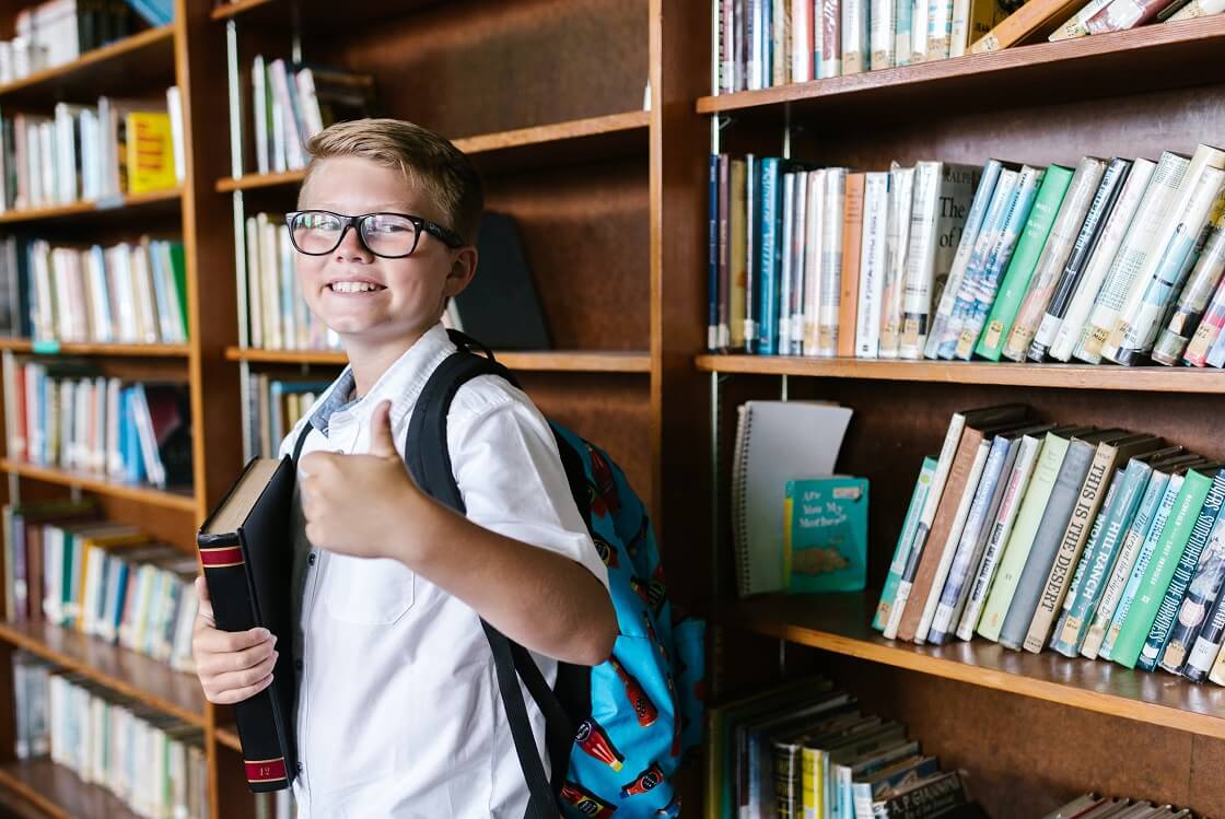 boy wearing glasses and backpack holding book giving thumbs up in library