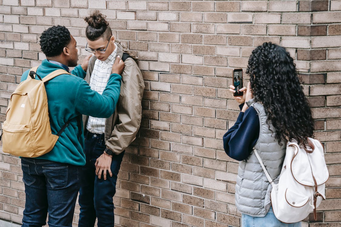 Student bullies another by pushing them against brick wall while another takes pictures with cell phone