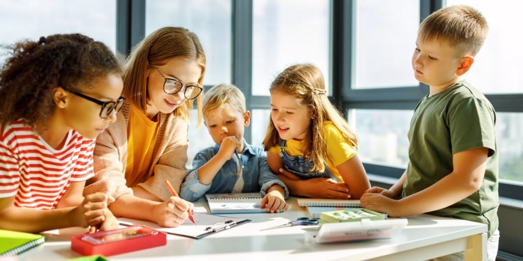 Happy female tutor and optimistic children smiling and discussing topic of lesson while gathering around table at school