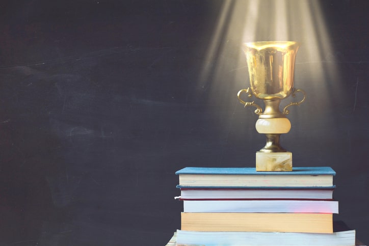 Golden trophy on pile of books, against blackboard, with sun rays over trophy