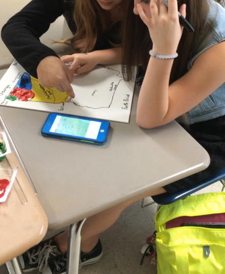 Two students work on a poster of a map. One is coloring while the other references an app on her mobile phone.