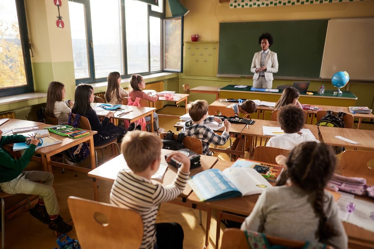 Rear view of large group of school kids listening to female teacher in class at elementary school