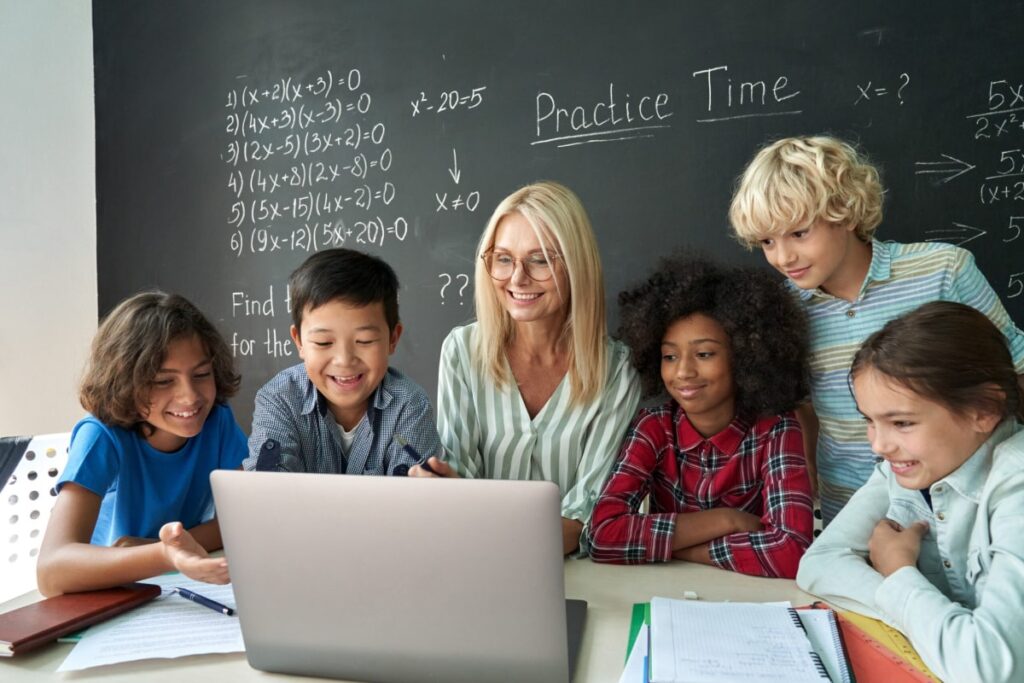 happy diverse junior school students gathered at teacher's desk looking at laptop