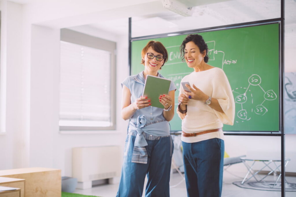 two female teachers standing in front of chalkboard in empty classroom with tablet