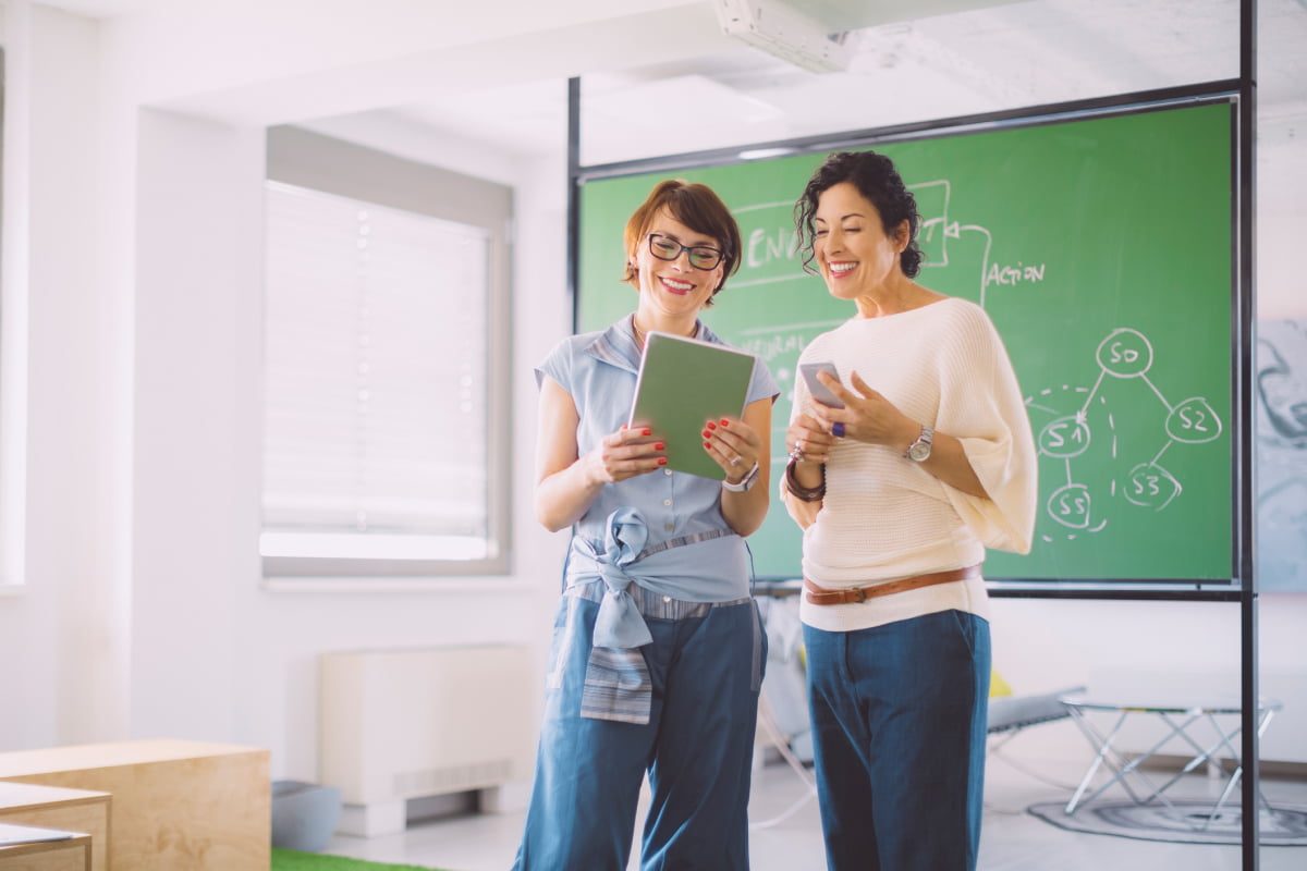 two female teachers standing in front of chalkboard in empty classroom with tablet
