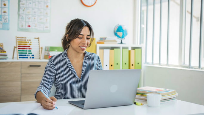 Teacher in classroom at desk using laptop talking with students on video