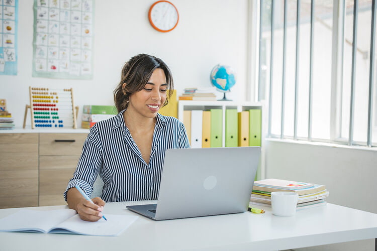 Teacher in classroom at desk using laptop while writing in notepad
