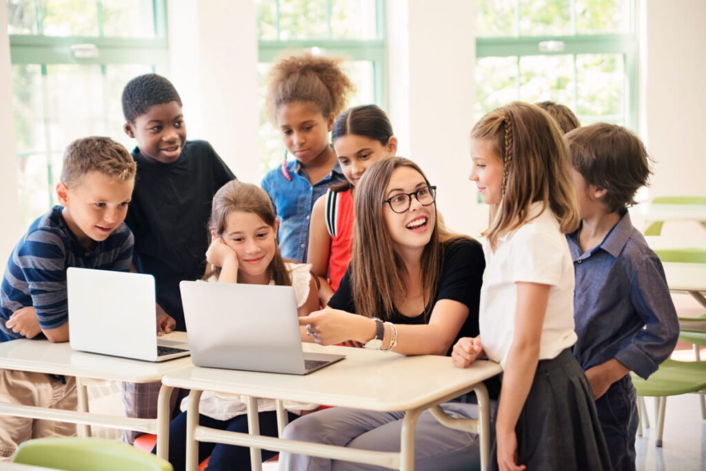Elementary school classroom teacher on laptop while students gather around